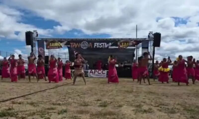 Group of dancers in pink skirts performing on a sandy festival ground in front of a stage with a banner overhead reading Festival and related logos from the event stage area.