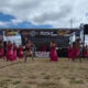 Group of dancers in pink skirts performing on a sandy festival ground in front of a stage with a banner overhead reading Festival and related logos from the event stage area.