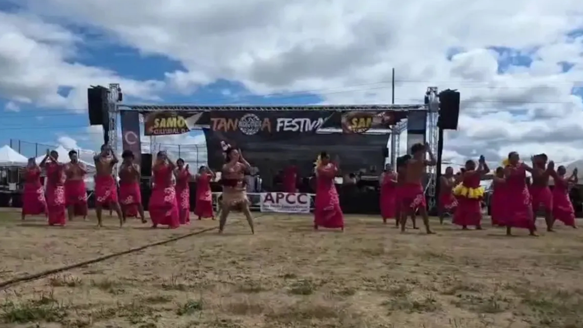 Group of dancers in pink skirts performing on a sandy festival ground in front of a stage with a banner overhead reading Festival and related logos from the event stage area.