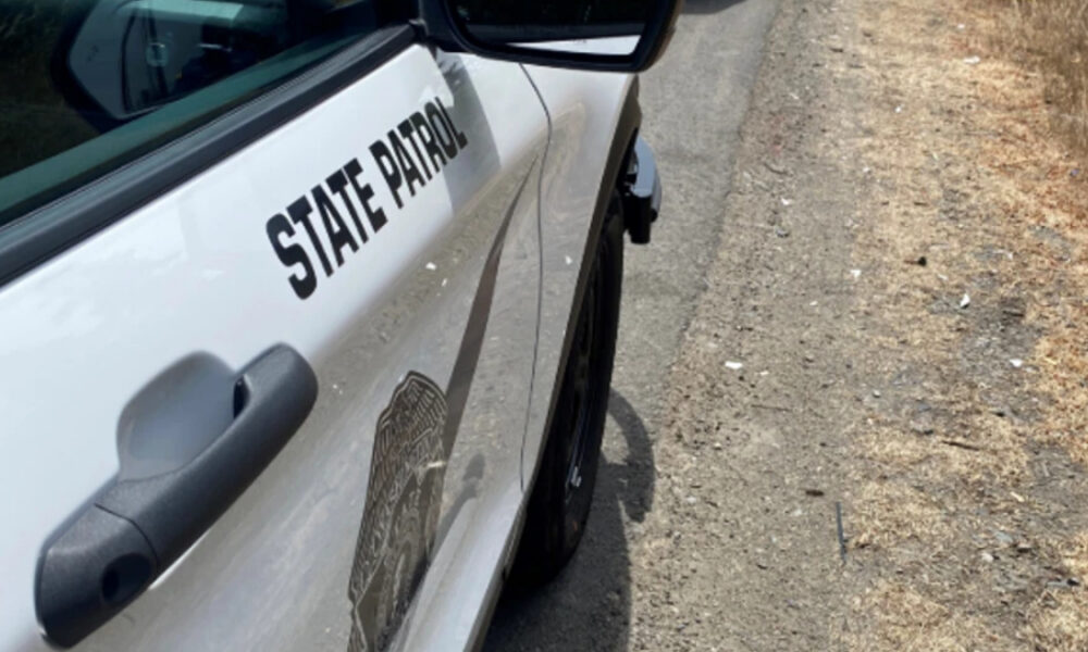 White state patrol police car parked on a dirt roadside, showing 'STATE PATROL' lettering on the door and a badge emblem visible on the side.