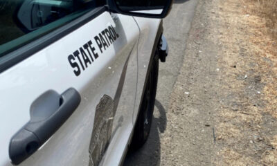 White state patrol police car parked on a dirt roadside, showing 'STATE PATROL' lettering on the door and a badge emblem visible on the side.