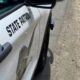 White state patrol police car parked on a dirt roadside, showing 'STATE PATROL' lettering on the door and a badge emblem visible on the side.