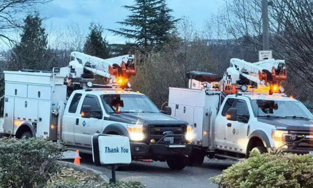 Two white utility trucks with bucket lifts parked side by side in a driveway, orange warning lights flashing on top, with a small 'Thank You' sign in the foreground.
