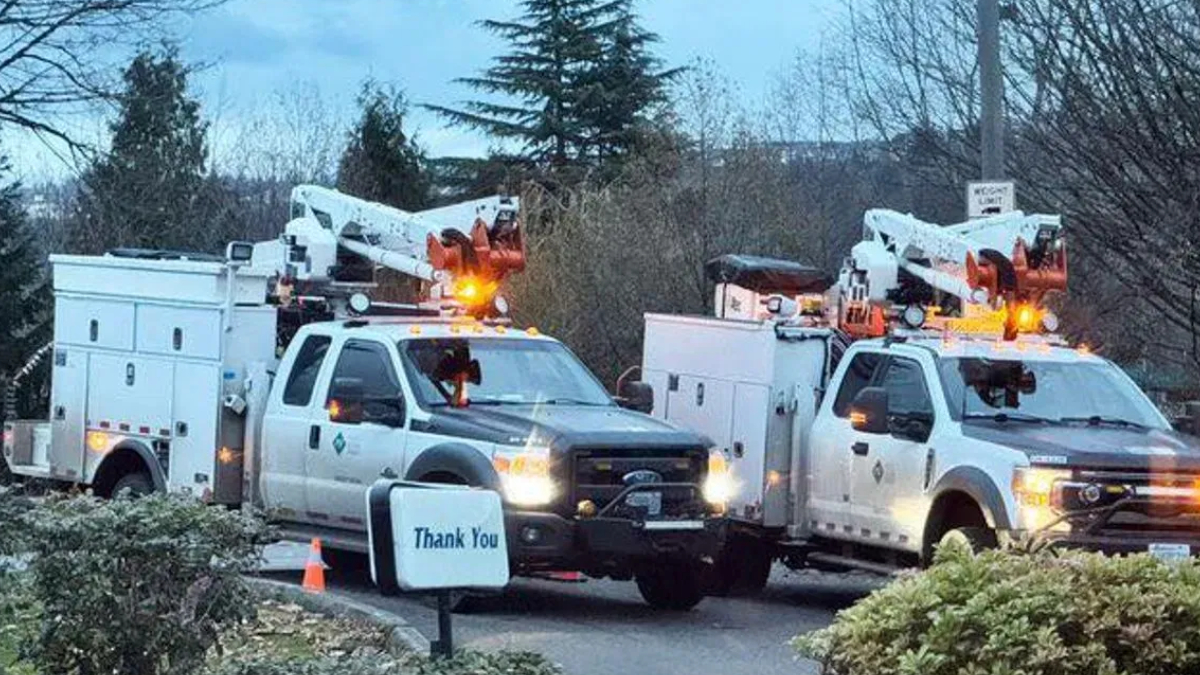 Two white utility trucks with bucket lifts parked side by side in a driveway, orange warning lights flashing on top, with a small 'Thank You' sign in the foreground.
