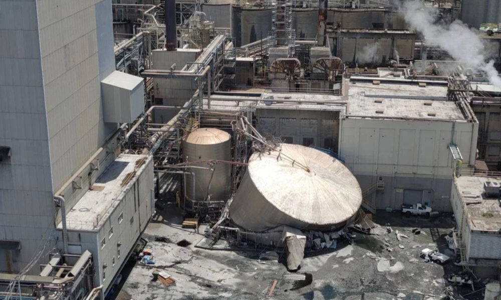 Industrial plant with a toppled circular metal storage tank, collapsed debris, and steaming pipes nearby.
