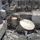 Industrial plant with a toppled circular metal storage tank, collapsed debris, and steaming pipes nearby.
