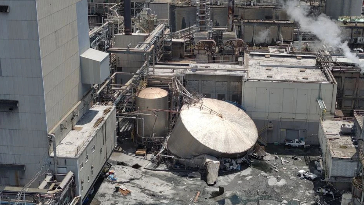 Industrial plant with a toppled circular metal storage tank, collapsed debris, and steaming pipes nearby.
