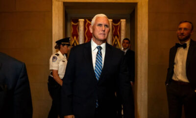 Older man in a dark suit with a blue striped tie walks down a marble hallway, staff visible in the background.