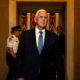 Older man in a dark suit with a blue striped tie walks down a marble hallway, staff visible in the background.