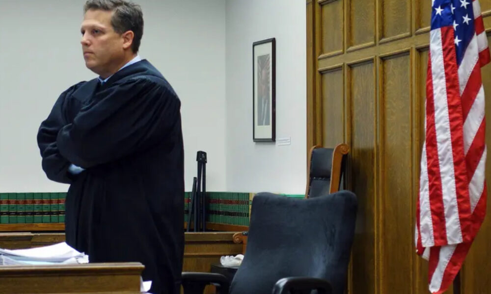 A judge in a black robe stands with arms crossed in a courtroom, papers on the desk nearby and an American flag to the right.