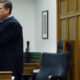 A judge in a black robe stands with arms crossed in a courtroom, papers on the desk nearby and an American flag to the right.