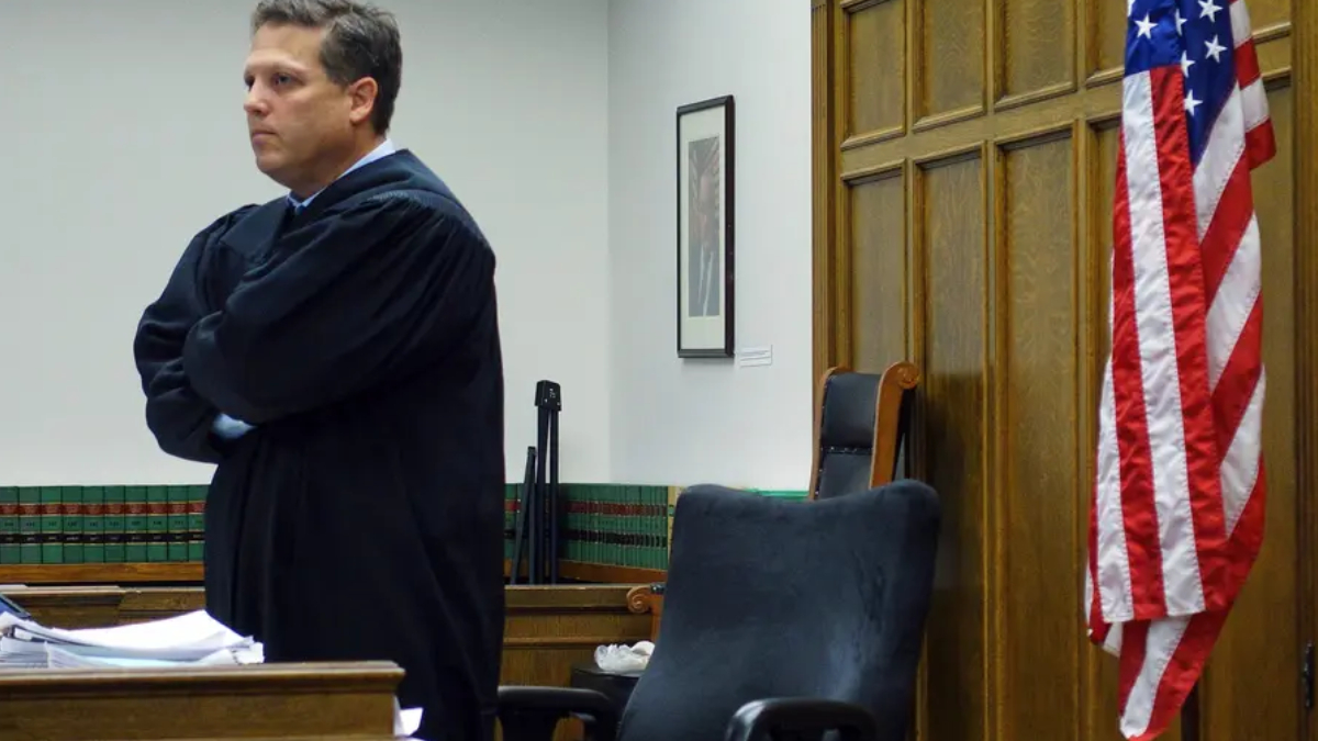 A judge in a black robe stands with arms crossed in a courtroom, papers on the desk nearby and an American flag to the right.