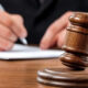 Wooden judge's gavel resting on a sound block with a person signing a document in the background.