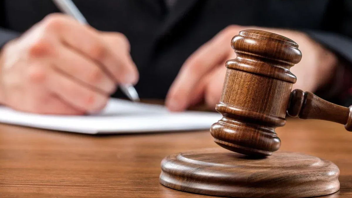 Wooden judge's gavel resting on a sound block with a person signing a document in the background.