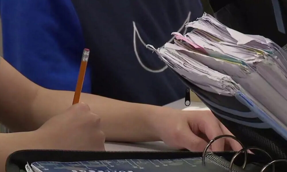 Student writes with a yellow pencil at a desk, papers and notebooks stacked nearby in a classroom setting.