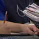 Student writes with a yellow pencil at a desk, papers and notebooks stacked nearby in a classroom setting.