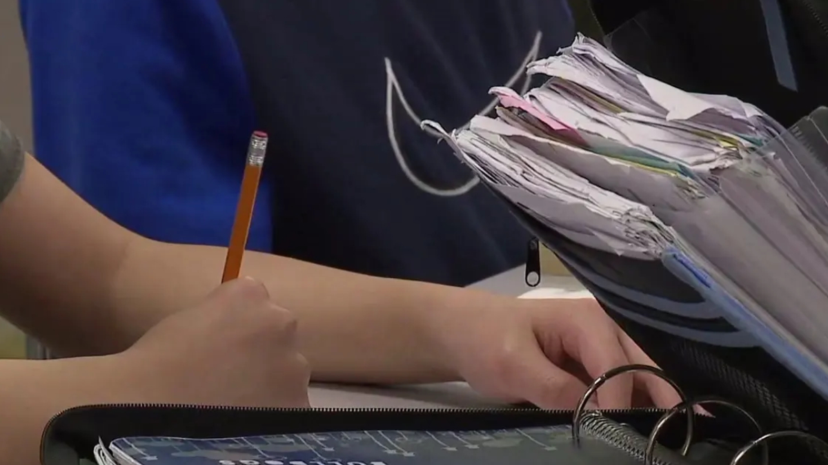 Student writes with a yellow pencil at a desk, papers and notebooks stacked nearby in a classroom setting.