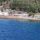 Shoreline town by a calm blue lake: white building near the water, parked cars, and a tree-lined backdrop with power lines overhead.”,