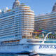 Large white cruise ship with colorful water slides on top, labeled Icon of the Seas, sailing on calm blue water with a small tender nearby