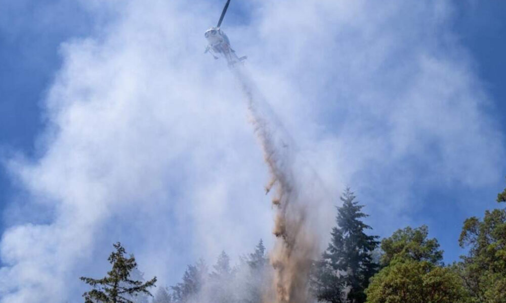 Firefighting helicopter releasing a long plume of retardant over a forest hillside with smoke and blue sky above.