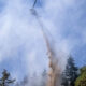 Firefighting helicopter releasing a long plume of retardant over a forest hillside with smoke and blue sky above.