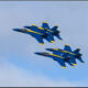 Two Blue Angels fighter jets flying in tight formation against a blue sky with light clouds.