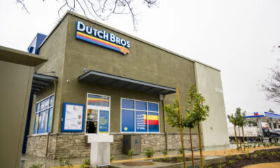Exterior of a Dutch Bros Coffee shop with olive-green walls, blue logo sign, and stone base accents.