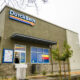 Exterior of a Dutch Bros Coffee shop with olive-green walls, blue logo sign, and stone base accents.