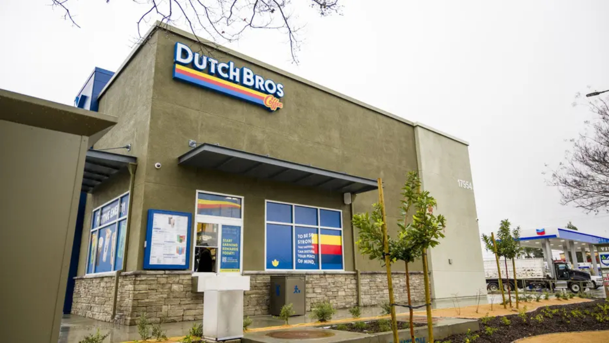 Exterior of a Dutch Bros Coffee shop with olive-green walls, blue logo sign, and stone base accents.