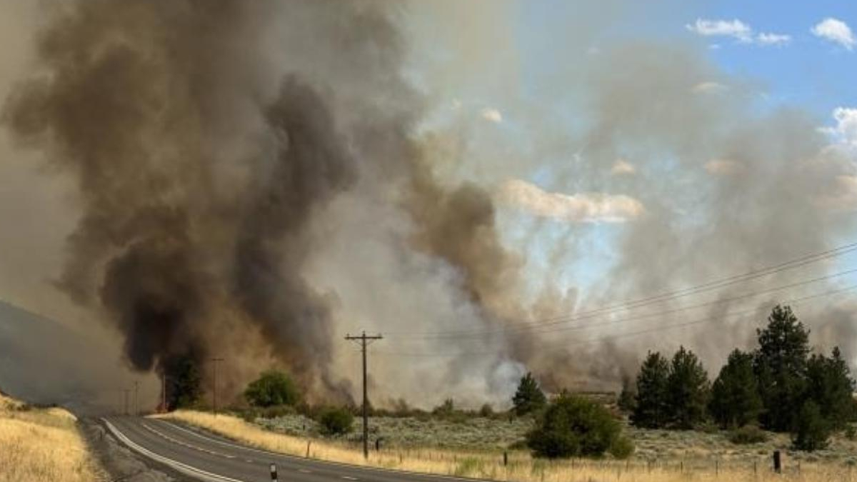 Rural highway with a massive wildfire to the left, thick dark smoke billowing into the sky.