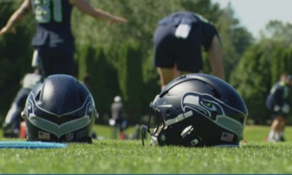 Two football helmets sit on a green practice field, with players in the blurred background behind them.