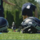 Two football helmets sit on a green practice field, with players in the blurred background behind them.