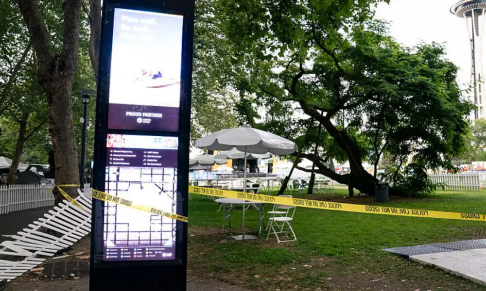 Tall digital information kiosk in a park, with yellow 'Police Line Do Not Cross' tape, white folding chairs, and white umbrellas under trees; Space Needle visible in the background.