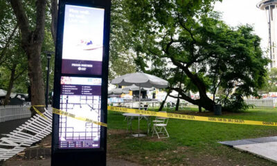 Tall digital information kiosk in a park, with yellow 'Police Line Do Not Cross' tape, white folding chairs, and white umbrellas under trees; Space Needle visible in the background.
