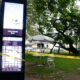 Tall digital information kiosk in a park, with yellow 'Police Line Do Not Cross' tape, white folding chairs, and white umbrellas under trees; Space Needle visible in the background.