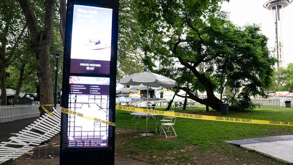 Tall digital information kiosk in a park, with yellow 'Police Line Do Not Cross' tape, white folding chairs, and white umbrellas under trees; Space Needle visible in the background.