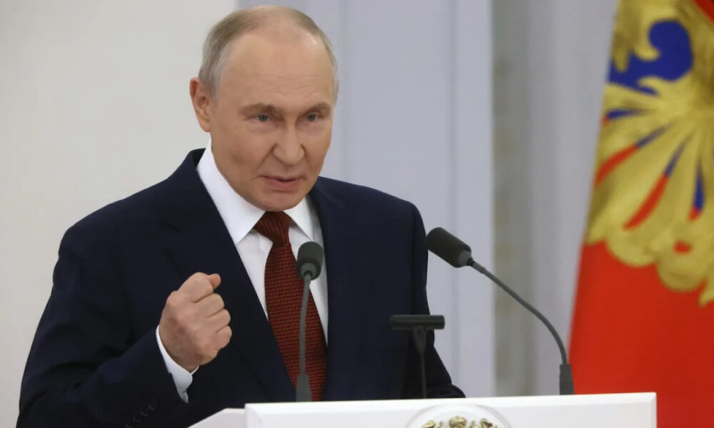 A man in a dark suit and red tie speaks at a podium with two microphones, his right fist raised in a gesture, against a backdrop with a decorative flag. (informative, describes action and setting)