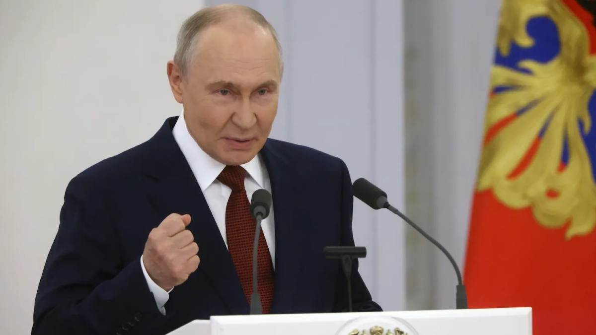 A man in a dark suit and red tie speaks at a podium with two microphones, his right fist raised in a gesture, against a backdrop with a decorative flag. (informative, describes action and setting)