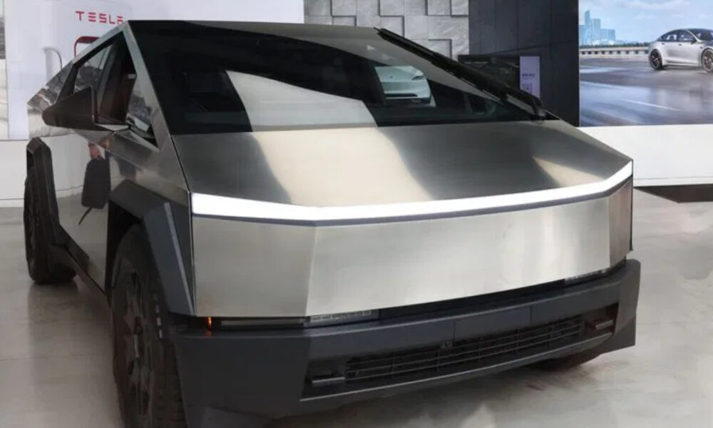 Front three-quarter view of a stainless steel Tesla Cybertruck on display in a showroom, featuring its angular panels and a thin LED light strip across the front.