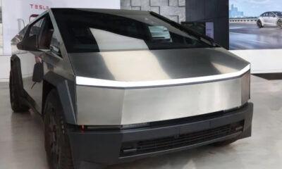 Front three-quarter view of a stainless steel Tesla Cybertruck on display in a showroom, featuring its angular panels and a thin LED light strip across the front.
