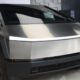 Front three-quarter view of a stainless steel Tesla Cybertruck on display in a showroom, featuring its angular panels and a thin LED light strip across the front.