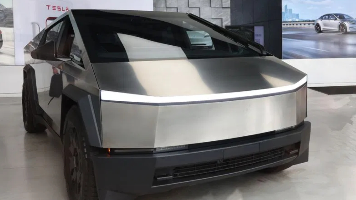 Front three-quarter view of a stainless steel Tesla Cybertruck on display in a showroom, featuring its angular panels and a thin LED light strip across the front.