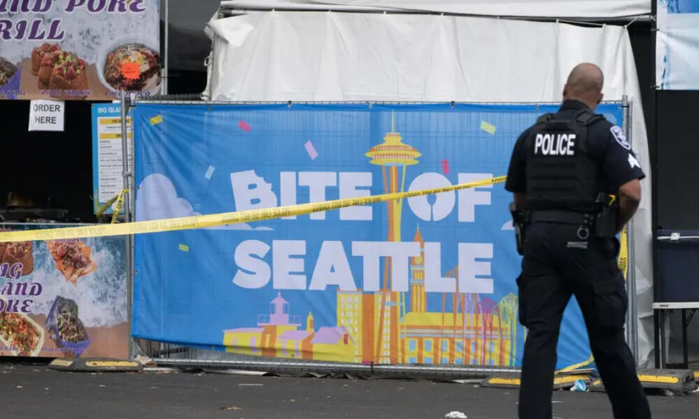 Police officer with back to camera guards a food festival booth; a blue banner reads 'BITE OF SEATTLE' with skyline illustrations and yellow caution tape across the scene.