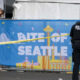 Police officer with back to camera guards a food festival booth; a blue banner reads 'BITE OF SEATTLE' with skyline illustrations and yellow caution tape across the scene.