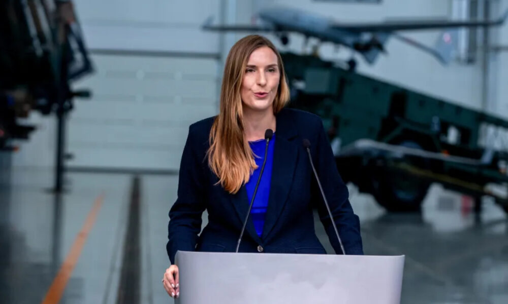 Woman in a dark blazer speaks at a podium with two microphones, in an aerospace exhibit audience-facing setup with a jet aircraft in the background.