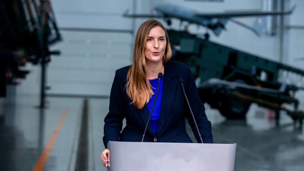 Woman in a dark blazer speaks at a podium with two microphones, in an aerospace exhibit audience-facing setup with a jet aircraft in the background.