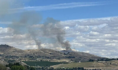 Smoke plumes rising from hills in a rural landscape, under a blue sky with scattered clouds.