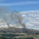 Smoke plumes rising from hills in a rural landscape, under a blue sky with scattered clouds.