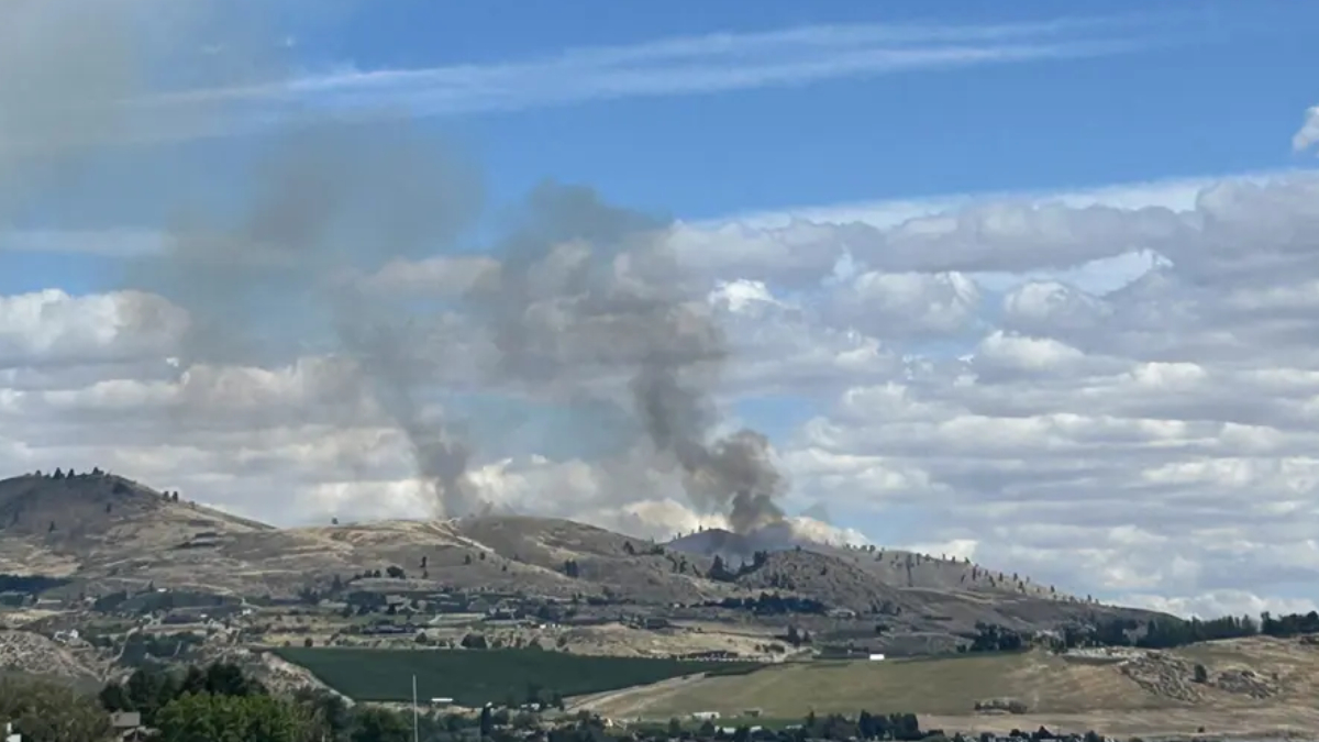 Smoke plumes rising from hills in a rural landscape, under a blue sky with scattered clouds.