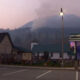 A small town street at dusk with a mountain backdrop, low buildings, a wooden fence, and a cafe edged by blue fairy lights.